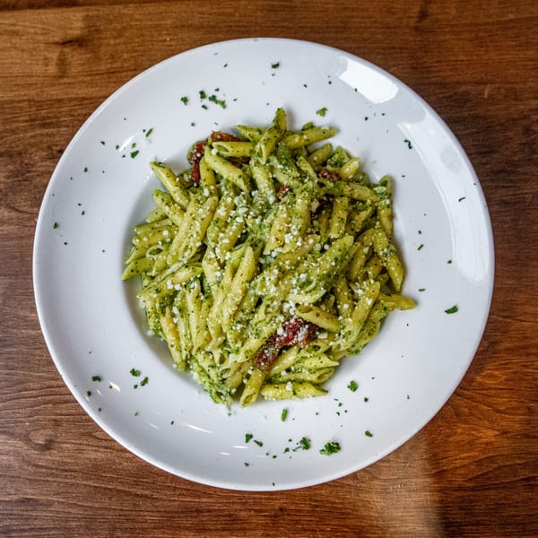 Cafe Pesto's penne pasta with green pesto, sun-dried tomatoes, and parmesan cheese on a white plate.