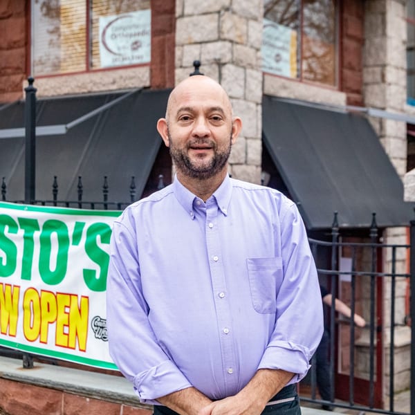 A smiling man in a purple shirt stands outside Cafe Pesto's, with a "Now Open" banner visible.