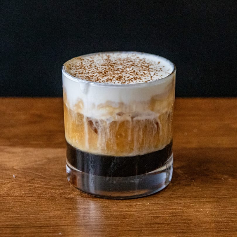 Layered drink with dark liquid, light brown liquid, and white foam topped with cinnamon on a wooden table.